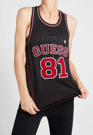 Kvinde iført sort, ærmeløs sportstrøje med røde og hvide bogstaver "GUESS 81", der holder i trøjens nederste kant.