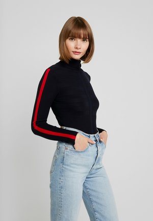 Navy turtleneck sweater with red side stripes, ribbed texture, form-fitting design. Paired with light blue high-waisted jeans.