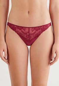 String en dentelle bordeaux avec un motif floral, présentant un dessus en mesh transparent et un tissu doux et extensible, avec des bords ornés de délicates finitions à la scallop.