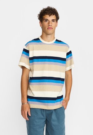 Jeune homme portant une chemise à manches courtes rayée beige, bleu, blanc et noir, et un pantalon bleu, debout avec une main dans la poche.