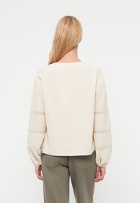 Vaalea beige puuvillainen pusero, jossa pyöreä pääntie, puffatut hihat ja herkät kirjailut hihoissa. Sileä rakenne, lyhyt malli.