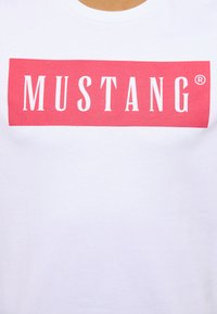 Biała bawełniana koszulka z czerwonym prostokątnym logo, na którym centralnie znajduje się napis "MUSTANG" w pogrubionych białych literach. Miękka faktura, standardowy okrągły dekolt.