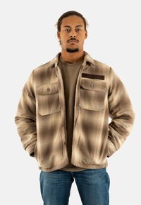 Chaqueta marrón de cuadros con dos bolsillos en el pecho, botones a presión y una etiqueta marrón. Llevada sobre una camisa beige y combinada con jeans azules.