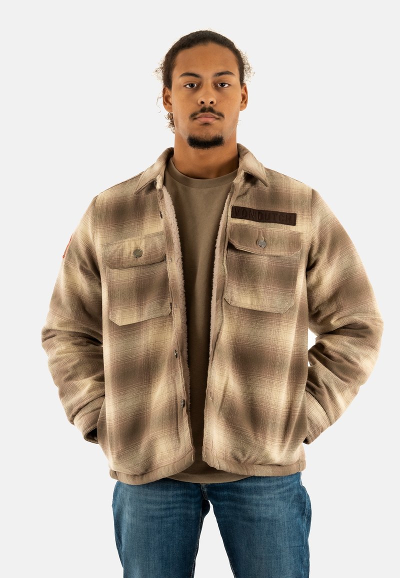 Chaqueta marrón de cuadros con dos bolsillos en el pecho, botones a presión y una etiqueta marrón. Llevada sobre una camisa beige y combinada con jeans azules.