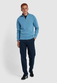 Pullover azzurro chiaro con collo a zip e polsini a costine, abbinato a pantaloni blu navy. Il modello è in piedi contro uno sfondo neutro, con le mani in tasca.