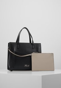 Černá kožená kabelka s texturovaným povrchem, dvojitými uchy a odnímatelnou béžovou peněženkou. Na přední straně je jemné logo.