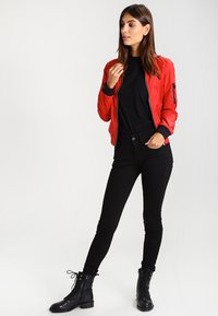 Chaqueta bomber roja con puños acanalados negros, usada sobre una camiseta negra. Unos vaqueros skinny negros y botas de tobillo negras con cordones completan el look.