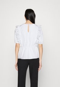 Weiße Bluse mit Puffärmeln, gerüschten Kanten und einem Lochmuster. Verfügt über eine Schlüssellochöffnung hinten und eine taillierte Passform.