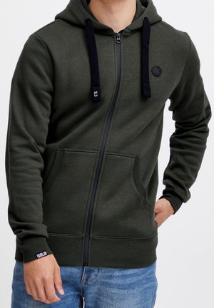 Homme portant un sweat à capuche olive à fermeture éclair avec cordons noirs, poches avant, et jean bleu, main posée sur la hanche et le côté.