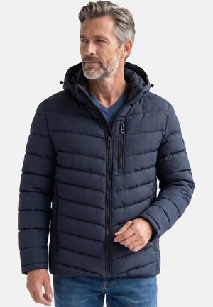 Mann mit grauen Haaren und Bart, der eine marineblaue gesteppte Kapuzenjacke und blaue Jeans trägt, schaut leicht zur Seite.