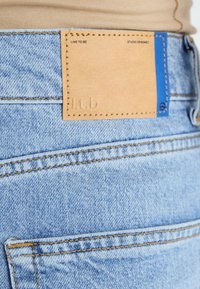 Light blue denim jeans with a tan leather patch on the waistband featuring embossed text, orange stitching, and a small blue accent.