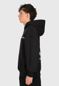 Giovane uomo con capelli ricci che indossa un hoodie nero, in piedi di profilo contro uno sfondo grigio chiaro uniforme.