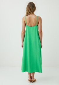Eine Frau mit langen Haaren steht mit dem Rücken zu uns und trägt ein lebhaft grünes ärmelloses Kleid, das bis zu ihren Knöcheln reicht, in einer minimalistischen weißen Umgebung.