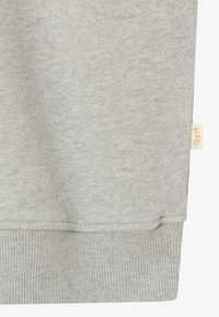 Jasnoszara bluza z dzianinowym ściągaczem u dołu i małą beżową metką po boku, na której częściowo widoczny jest tekst.