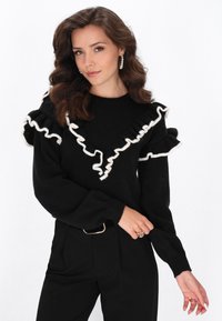 Maglione nero con dettagli di ruffle bianchi lungo il collo e le maniche. Abbinato a pantaloni neri a vita alta e una cintura con fibbia argentata.