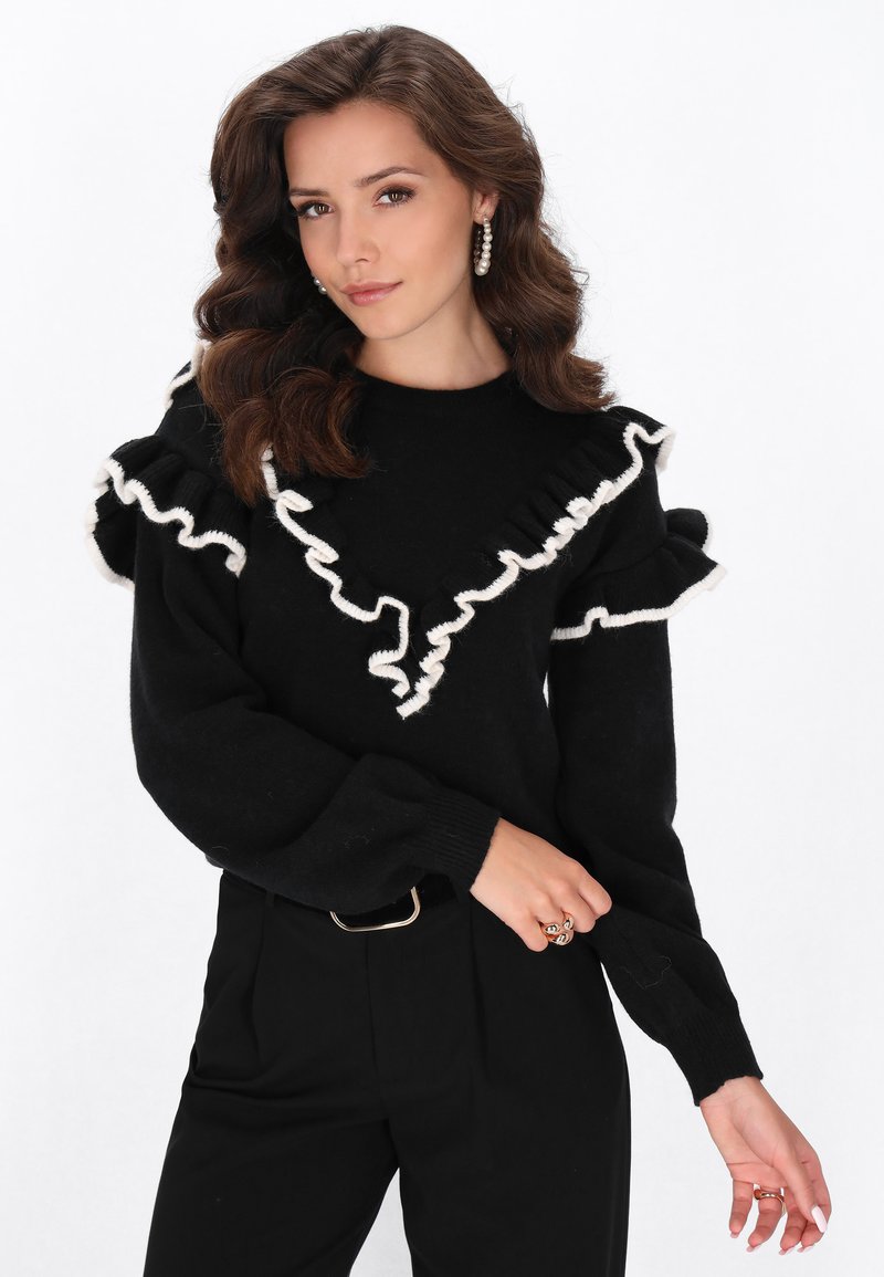 Maglione nero con dettagli di ruffle bianchi lungo il collo e le maniche. Abbinato a pantaloni neri a vita alta e una cintura con fibbia argentata.