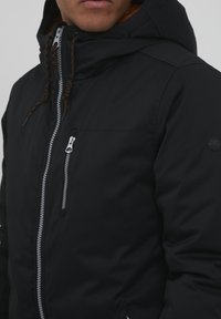 Black zip-up jacket with a hood, featuring contrasting orange lining, textured drawstrings, and a zippered chest pocket.