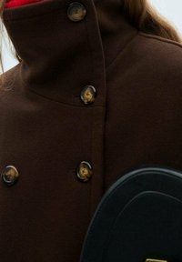 Cappotto di lana marrone con colletto alto, caratterizzato da grandi bottoni rotondi in tartaruga e una fodera interna rossa. Borsa nera visibile nell'inquadratura.