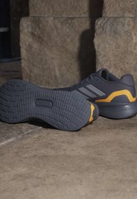 Chaussures de sport gris foncé avec des accents jaunes et des semelles en caoutchouc texturées, placées sur un sol en pierre devant un mur en pierre.