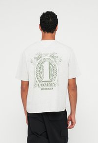 Grå bomull t-skjorte med kort erm. Baksiden har grønt grafisk design med "1" og teksten "TOMMY JEANS", omgitt av dekorative elementer.