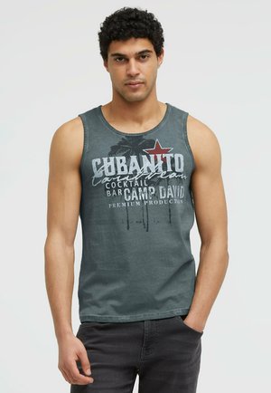 Mann mit kurzen lockigen dunklen Haaren, der ein ärmelloses graues Tanktop mit gedrucktem Text und schwarzen Hosen trägt, steht mit einer Hand in der Tasche.