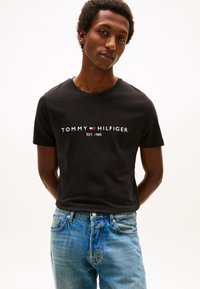 Svart t-skjorte i bomull med "TOMMY HILFIGER"-logo, rund hals, korte ermer og jevn tekstur, kombinert med blå jeans.