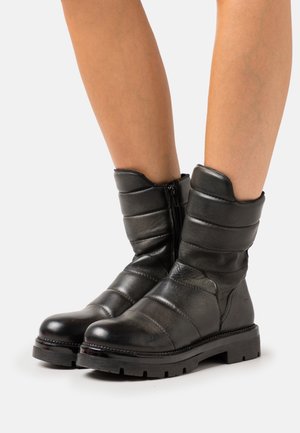 Jambes portant des bottes en cuir noir matelassé mi-mollet avec des fermetures éclair latérales, debout sur une surface blanche.