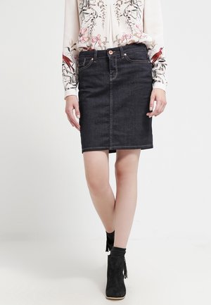 Kvinde iført en mørk denim knælang nederdel, blomsterprintet langærmet bluse, sorte ankelstøvler, stående med benene krydset.