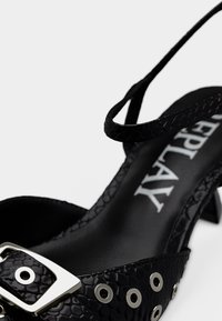 Μαύρο πέδιλο με τακούνι και υφή στην επιφάνεια, με τετράγωνη ασημένια αγκράφα και κυκλικά διακοσμητικά δακτυλίδια· περιλαμβάνεται λεπτή λουρίδα στο αστράγαλο.