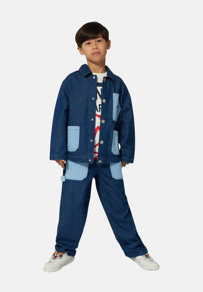 Ensemble en jean comprenant une veste et un pantalon de bleu foncé et clair, avec de grandes poches à rabat et des boutons en métal. Comprend un t-shirt graphique en dessous.