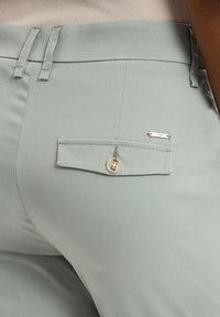 Hellgrüne Hose mit glatter Textur, ausgestattet mit einer Gesäßtasche mit Knopfverschluss und einem kleinen Metall-Label.