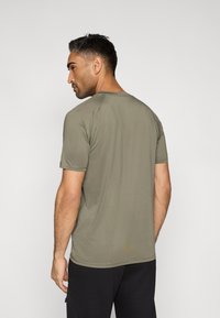 Oliwkowo-zielona koszulka z krótkim rękawem i okrągłym dekoltem, o gładkiej fakturze i subtelnych detalach przeszyć, noszona z czarnymi szortami.