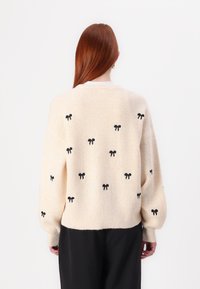 Pull en tricot beige doux, avec des nœuds brodés noirs, une texture côtelée et une coupe ample avec des emmanchures tombantes.