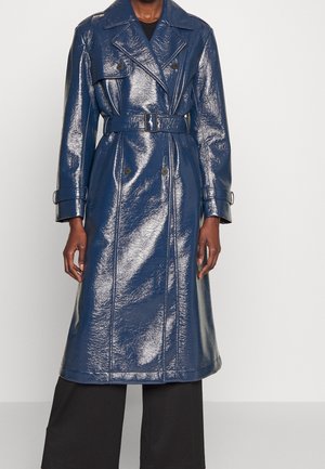 Trench-coat bleu marine brillant ceinturé avec larges revers et détails de boutons porté sur un pantalon noir à jambes larges sur une personne debout.