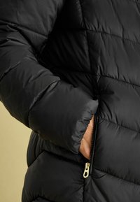 Melns pūkains mētelis ar izšūtu dizainu un gludu tekstūru, ar rāvējslēdzēja sānu kabatu un cieši pieguļošu aproci.