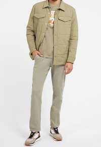 Polstret olivgrøn jakke med trykknapper og to brystlommer, kombineret med en beige t-shirt med et farverigt motiv og lyse bukser.