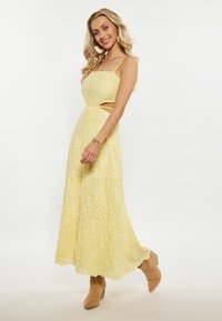 Rochie maxi galbenă, fără mâneci, cu design cu decupaje și modele brodate; tiv cu margini ondulate; purtată cu botine bej până la gleznă.
