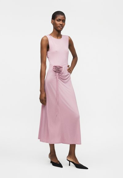 Γυναίκα με ροζ midi φόρεμα χωρίς μανίκια και λεπτομέρεια ρόζτας στη μέση, συνδυασμένο με μαύρες μυτερές γόβες, στέκεται μπροστά σε λευκό φόντο.