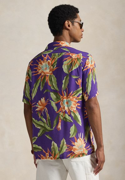 Mężczyzna w okularach przeciwsłonecznych i fioletowej koszuli z krótkim rękawem z dużym pomarańczowo-zielonym kwiatowym wzorem, w białych spodniach, stojący bokiem.