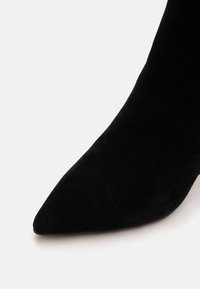 Gros plan d'une pointe de chaussure en daim noir à bout pointu sur un fond blanc uni.