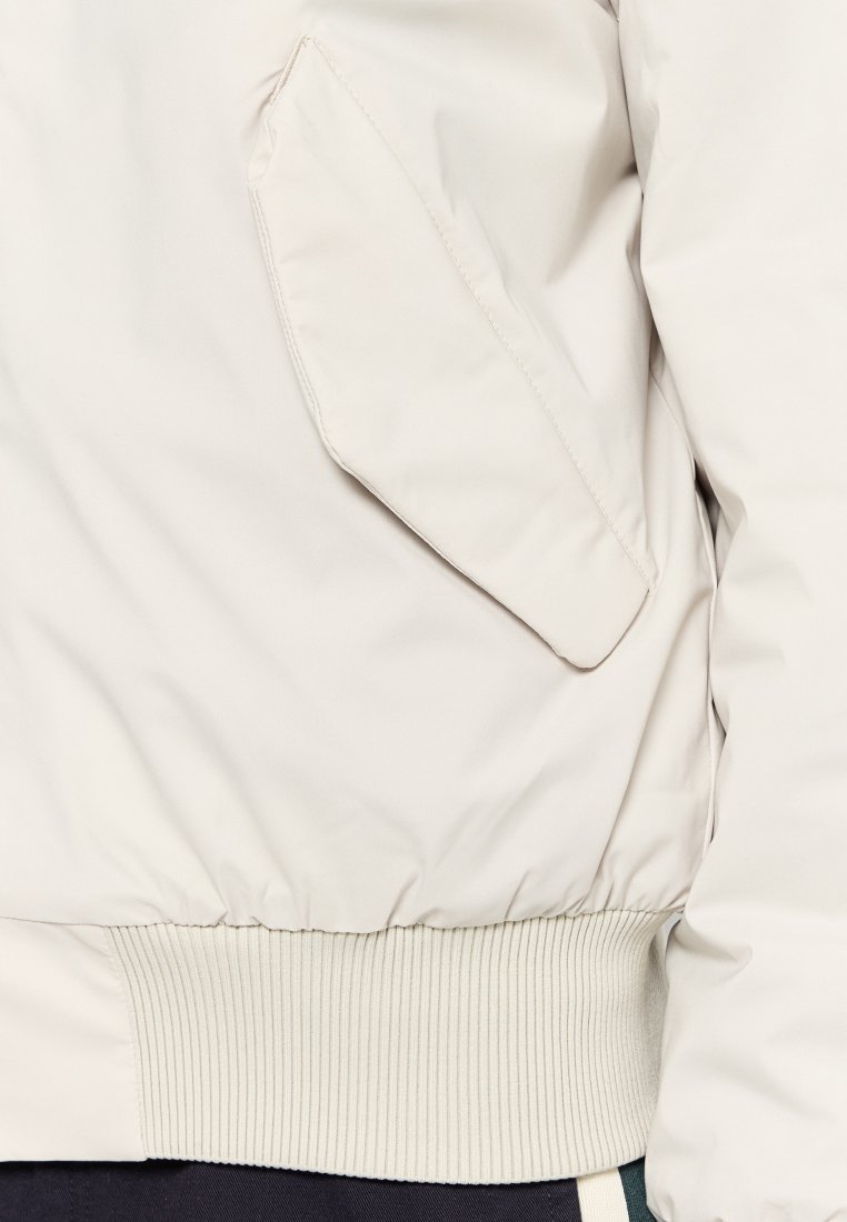 Sezione di giacca imbottita beige che mostra una tasca a pattina inclinata e un elastico a coste in vita.