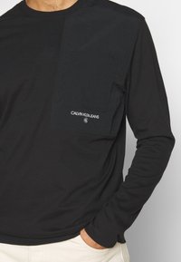 Černá dlouhá rukávová košile s texturovanou kapsou na levé hrudi, která má bílou vyšívanou značku. Měkká tkanina s volným střihem.