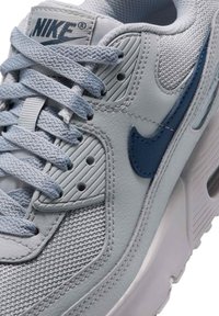 Κοντινή λήψη ενός ανοιχτού γκρι αθλητικού παπουτσιού Nike με μπλε λογότυπο, υφασμένο πλέγμα, δερμάτινα πάνελ και πλεκτά κορδόνια.