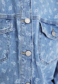 Denim srajca v svetlo modri barvi z bledim cvetličnim vzorcem, z enim srebrnim pritrdilnim gumbom, dvema žepoma na prsih in vidnimi šivi.