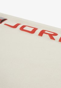 Ύφασμα σε κρεμ απόχρωση με ανάγλυκο κόκκινο κείμενο "JORDAN". Διαθέτει απαλό υφή και διακριτικό σχέδιο. Καθαρός σχεδιασμός, χωρίς επιπλέον διακοσμητικά.