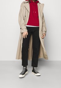Beige trenchcoat över en röd hoodie, svarta jeans och svarta högstövlar med vita sulor. Outfiten har en lagrad look med en avslappnad passform.