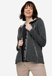 Huvjacka i mörkblått med en prickig textur, med en dragkedja fram, två sidofickor och kontrasterande ljusa dragkedjedetaljer.