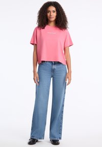 Růžové zkrácené tričko s krátkým rukávem s logem "GUESS JEANS". V kombinaci s modrými džíny s širokými nohavicemi a černými botami. Hladká textura.