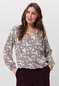 Beige blouse met een bruin geometrisch patroon, V-hals, lange mouwen met geplooide manchetten en een losse pasvorm, gecombineerd met donkerpaarse broek.