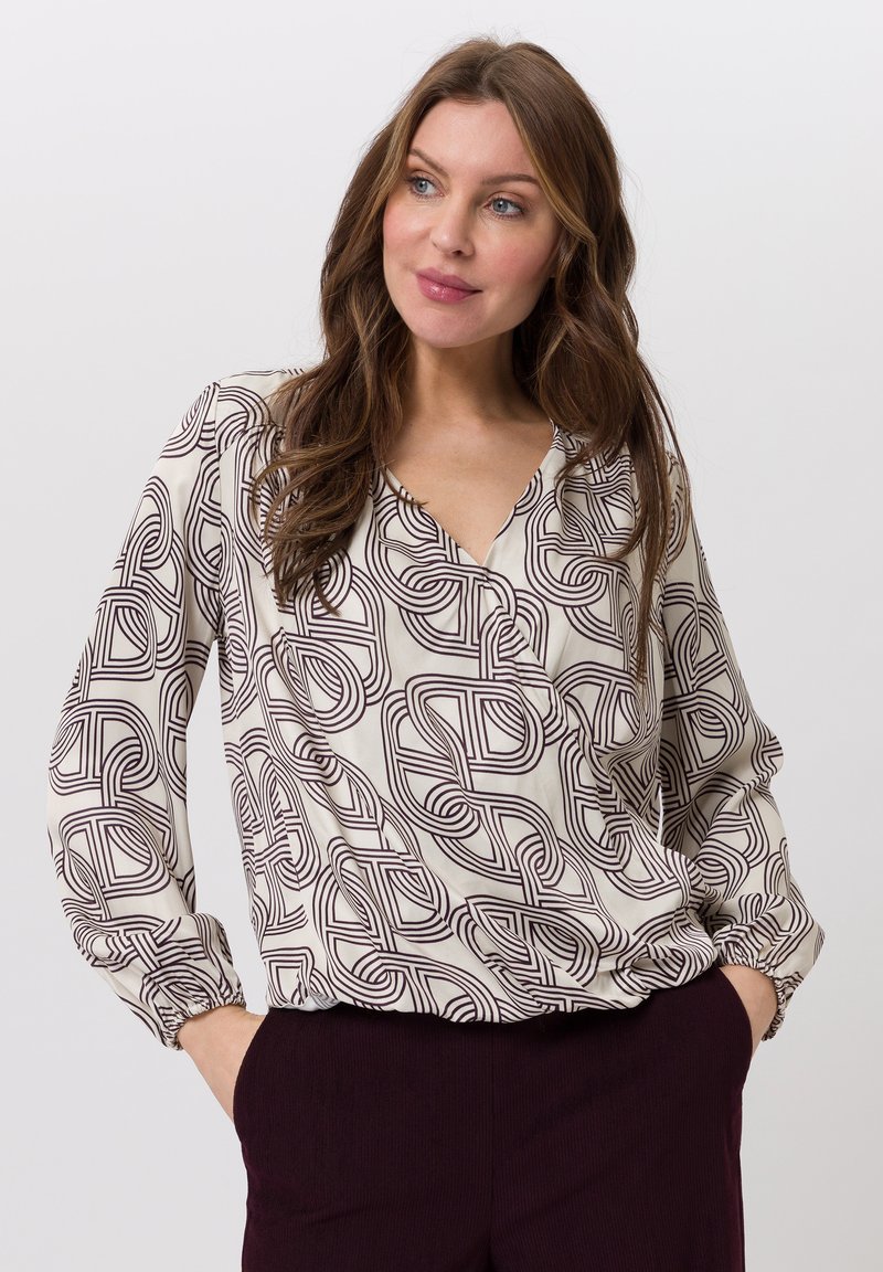 Beige blouse met een bruin geometrisch patroon, V-hals, lange mouwen met geplooide manchetten en een losse pasvorm, gecombineerd met donkerpaarse broek.